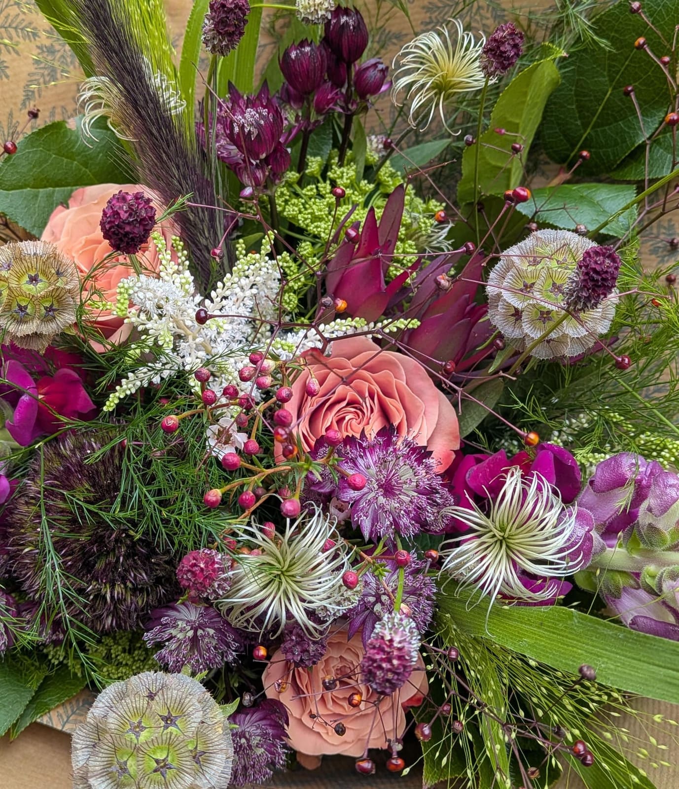 A wild hand-tied bouquet of peach roses, plum astrantia, clematis seedheads, scabiosa pods and sweet peas on patterned botanical paper