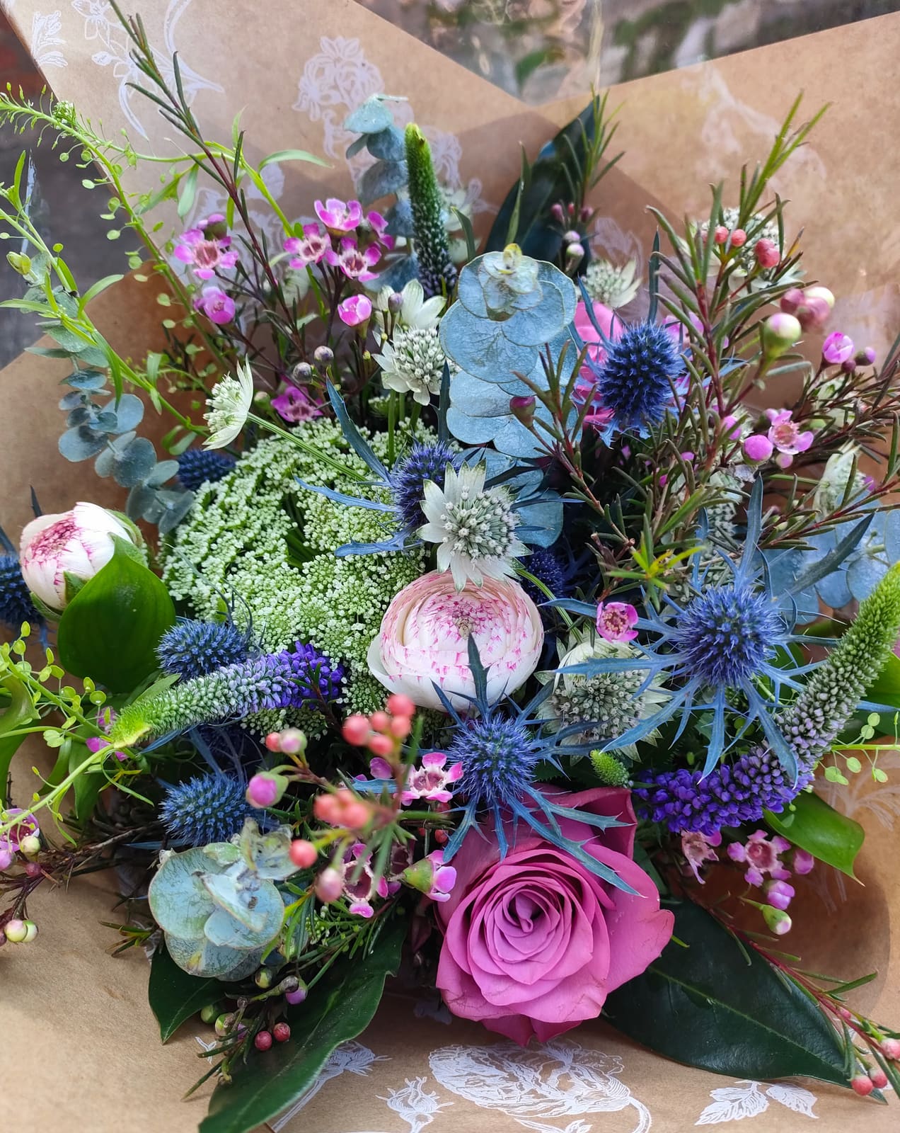 A hand-tied bouquet of pink rose, blue sea holly, waxflower and eucalyptus wrapped in botanical kraft paper