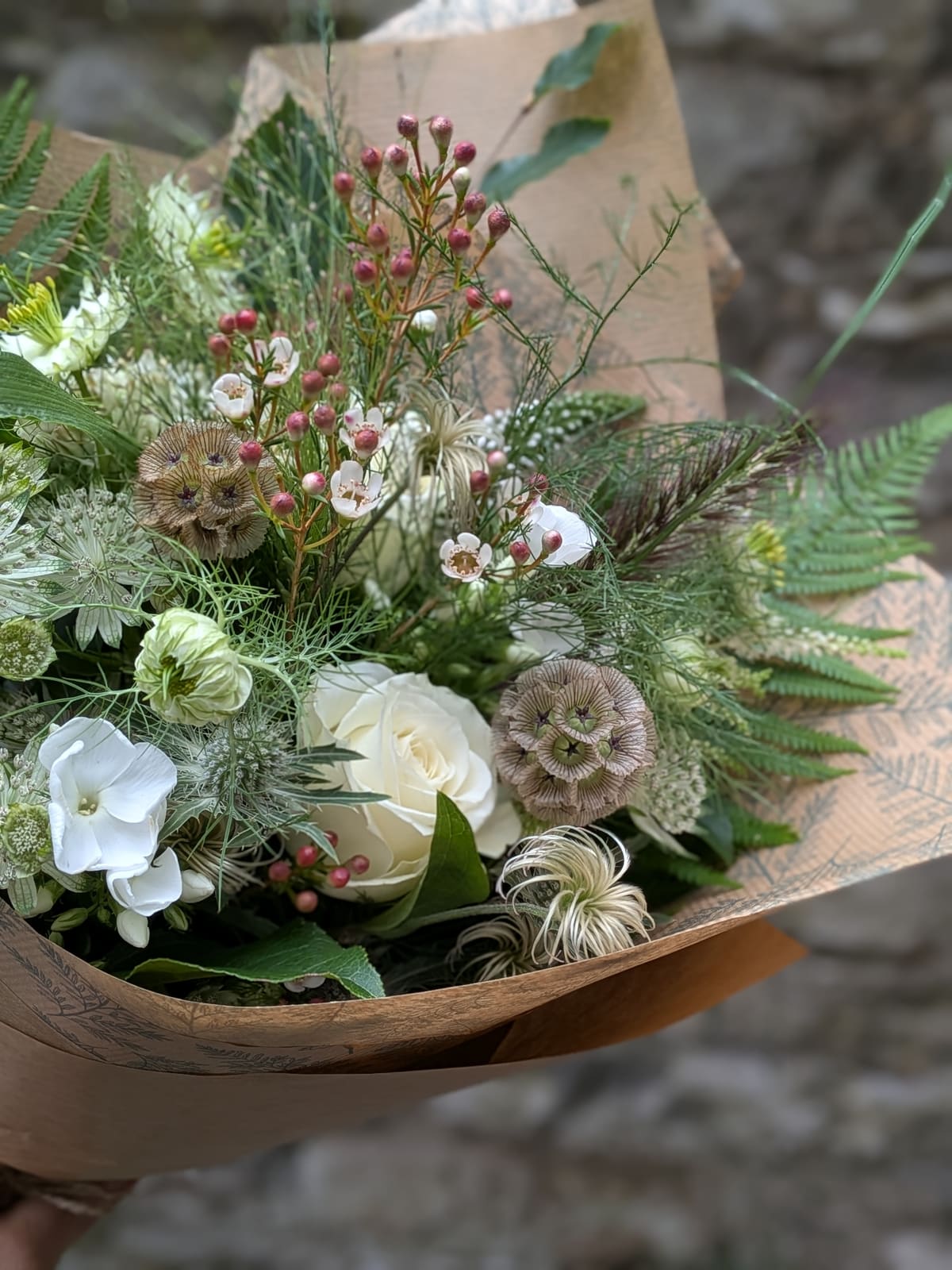 A natural white and green bridal-style bouquet of roses, waxflower, scabiosa seedheads and ferns wrapped in kraft paper