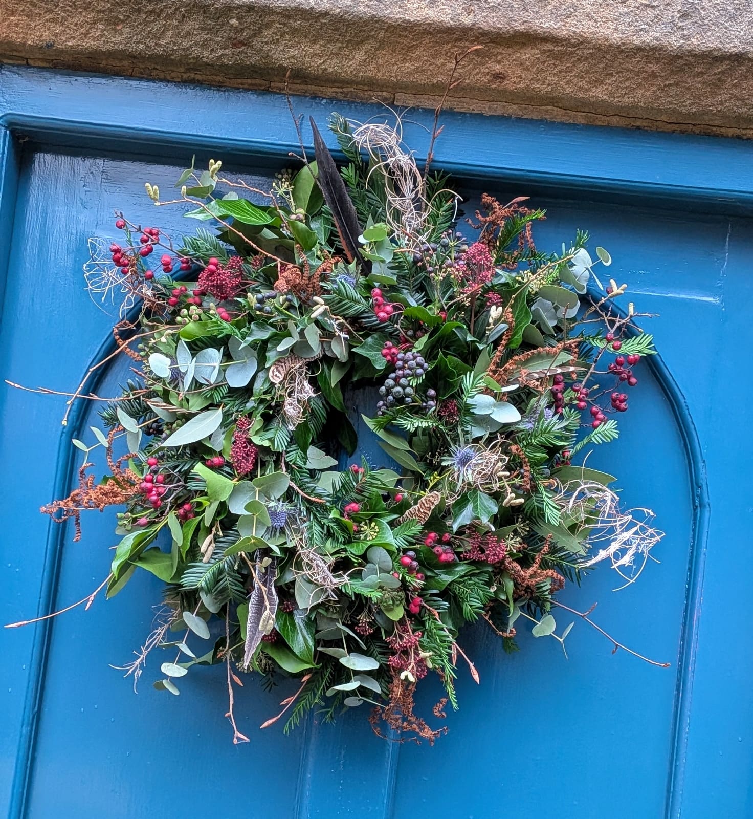 A foraged fresh Christmas wreath of yew, ivy, eucalyptus, berries and pheasant feathers on a blue front door