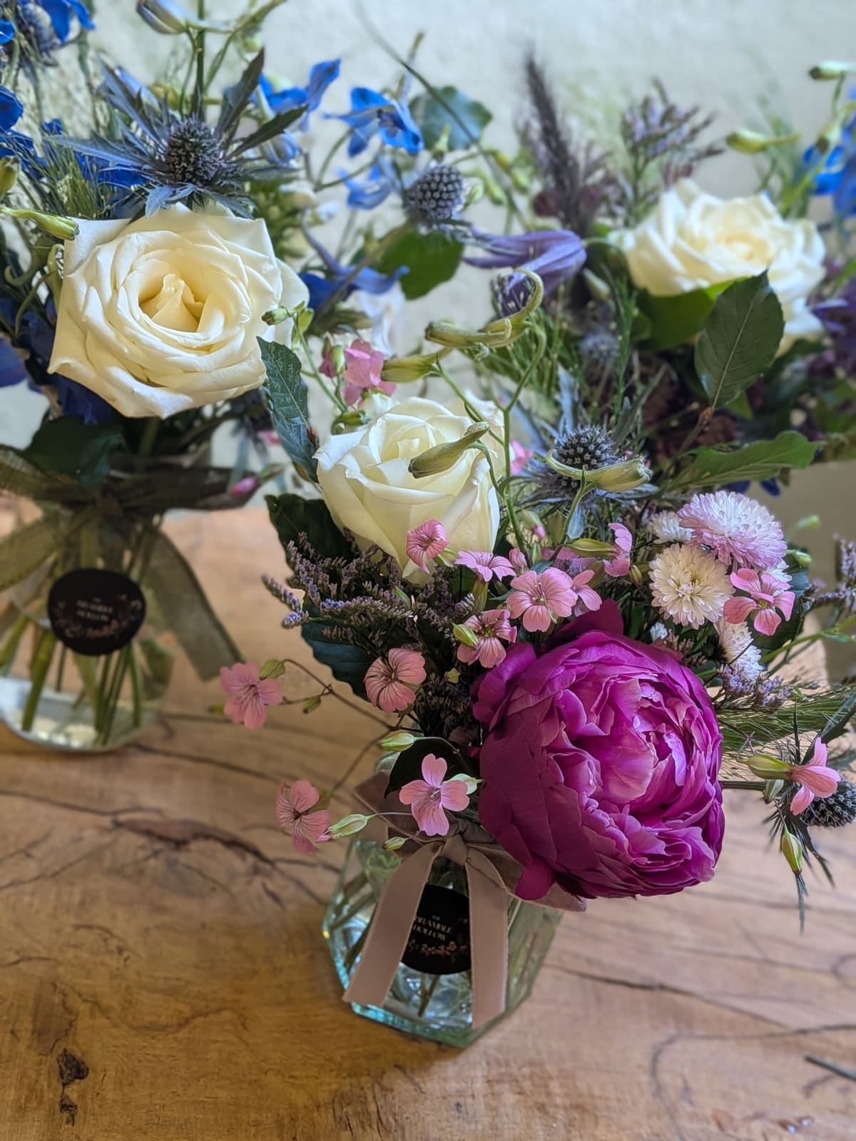 Little jar arrangements with a deep pink peony, white roses, blue delphinium and sea holly on a wooden counter