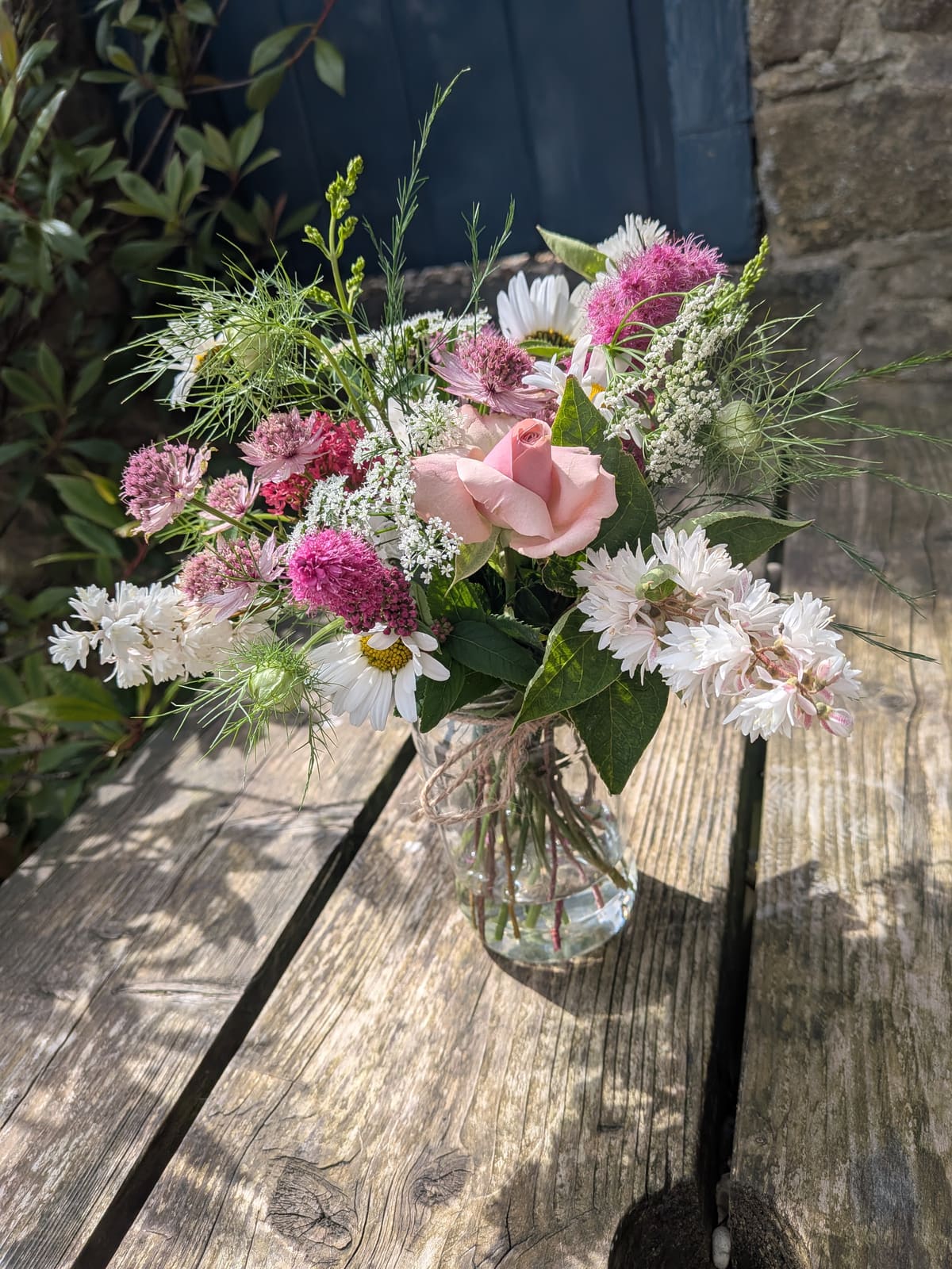 A summer posy of pink roses, daisies, astrantia and cow parsley in a glass jar on a garden bench